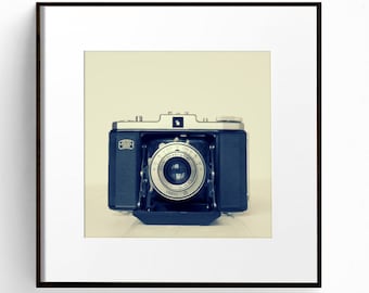 May include: A vintage black and silver camera with a round lens. The camera is in focus and the background is blurred.