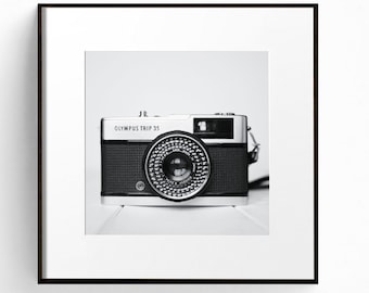 May include: A black and silver Olympus Trip 35 film camera with a black leather strap. The camera is sitting on a white surface.