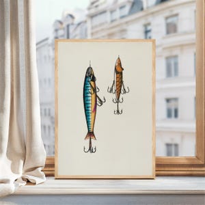 May include: A framed print featuring two vintage fishing lures. One lure is blue, green, and yellow, while the other is brown. Both lures have multiple hooks and are set against a cream-colored background.