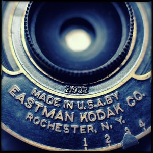 May include: Close-up of a vintage camera lens with the text "MADE IN U.S.A. BY EASTMAN KODAK CO. ROCHESTER, N.Y." and the number "21932" printed on the metal.