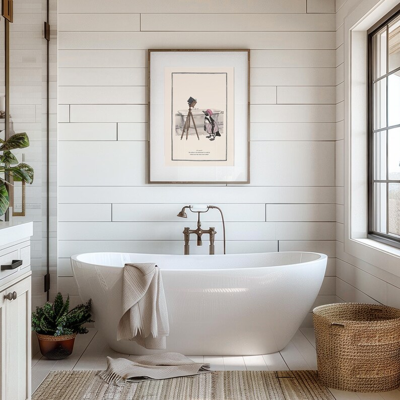 May include: A modern bathroom with a white, oval-shaped bathtub. A framed vintage print hangs above the tub. A woven basket and potted plant add natural elements. A beige towel is draped over the tub's edge. The walls are white.