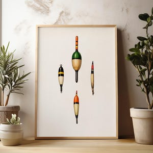 May include: A framed print featuring four vintage fishing bobbers. The bobbers are in various shapes and colors, including green, orange, black, and yellow. The print is displayed on a light-colored wall, with potted plants on either side.