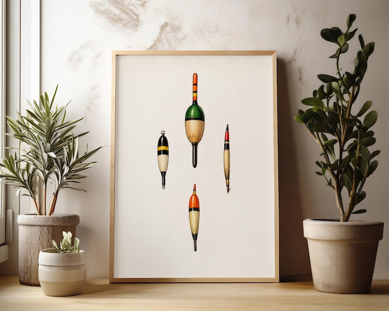 May include: A framed print featuring four vintage fishing bobbers. The bobbers are in various shapes and colors, including green, orange, black, and yellow. The print is displayed on a light-colored wall, with potted plants on either side.