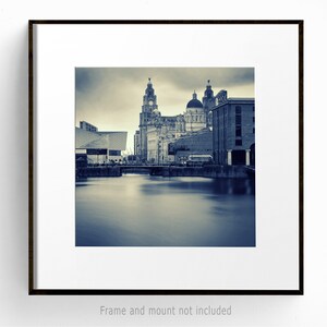 May include: A framed print of a cityscape in sepia tones. The image features a body of water in the foreground, with buildings and a clock tower in the background. The print is mounted in a white mat and a dark brown frame.