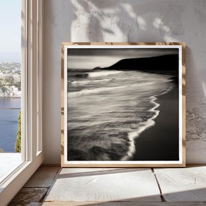 May include: A black and white photograph of a beach scene with waves crashing on the shore. The photo is framed in a light wood frame.