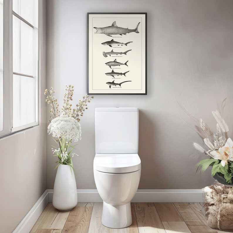 May include: A white toilet with a closed lid and a black framed print of different types of sharks hanging on the wall above it. There are white flowers in a white vase to the left of the toilet and dried flowers in a brown vase to the right of the toilet.