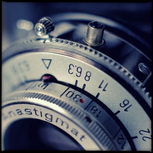 May include: Close-up of a vintage camera lens with a silver and black finish. The lens has a depth of field scale marked with numbers from 11 to 22 and a  'f: 8.3' marking. The lens is labelled 'anastigmat'.