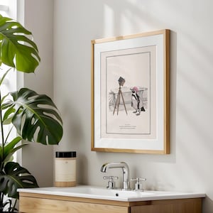 May include: Framed art print featuring a cartoon penguin in a striped outfit and pink hat, standing near a bathtub with a vintage camera. The print has a light wood frame and a white mat.