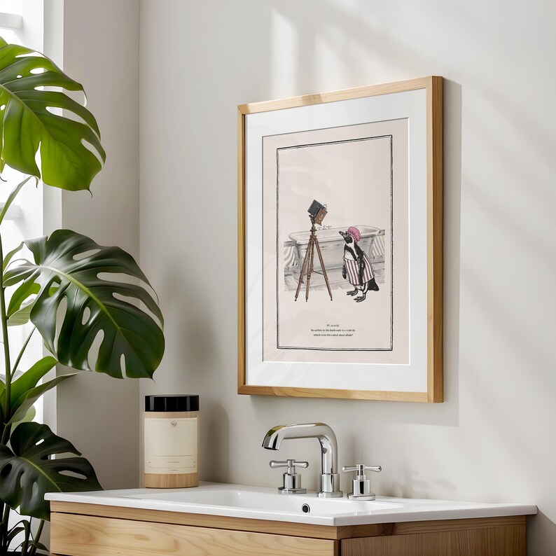 May include: Framed art print featuring a cartoon penguin in a striped outfit and pink hat, standing near a bathtub with a vintage camera. The print has a light wood frame and a white mat.