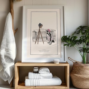 May include: Framed art print featuring a penguin in a striped outfit and a pink hat, posing with an antique camera in front of a bathtub. The print is in a white frame and rests on a wooden shelf with folded white towels.