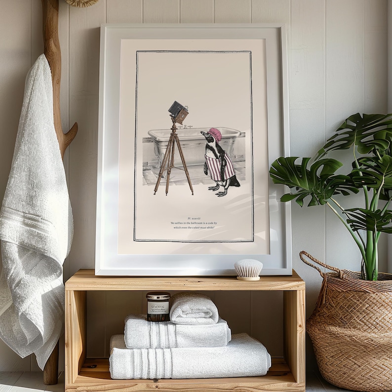 May include: Framed art print featuring a penguin in a striped outfit and a pink hat, posing with an antique camera in front of a bathtub. The print is in a white frame and rests on a wooden shelf with folded white towels.