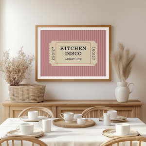 Puede incluir: Un cuadro enmarcado con el texto "KITCHEN DISCO ADMIT ONE" en un dise&ntilde;o de boleto. El fondo es un patr&oacute;n de rayas rosas. El cuadro est&aacute; colgado sobre una mesa de comedor con tazas y platos blancos.