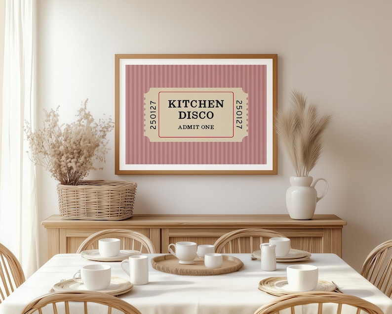 Puede incluir: Un cuadro enmarcado con el texto "KITCHEN DISCO ADMIT ONE" en un dise&ntilde;o de boleto. El fondo es un patr&oacute;n de rayas rosas. El cuadro est&aacute; colgado sobre una mesa de comedor con tazas y platos blancos.