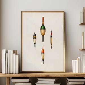 May include: A framed print featuring four vintage fishing bobbers. The bobbers are in various shapes and colors, including green, orange, yellow, and black. The print is displayed on a wooden shelf with books and a neutral wall background.