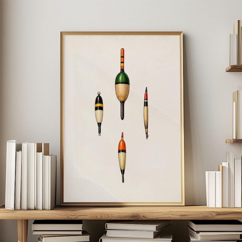 May include: A framed print featuring four vintage fishing bobbers. The bobbers are in various shapes and colors, including green, orange, yellow, and black. The print is displayed on a wooden shelf with books and a neutral wall background.