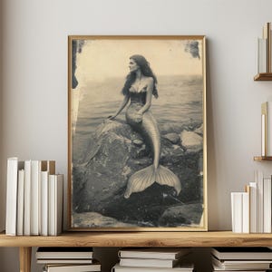 May include: A black and white vintage photograph of a mermaid sitting on a rock. The mermaid has long hair and is wearing a shell bra. Her tail is curled around the rock. The photo has a faded, antique look.