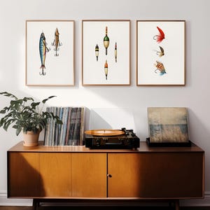 May include: Three framed art prints featuring fishing lures and bobbers. The prints are displayed above a mid-century modern style wooden cabinet with a record player, vinyl records, and a potted plant.