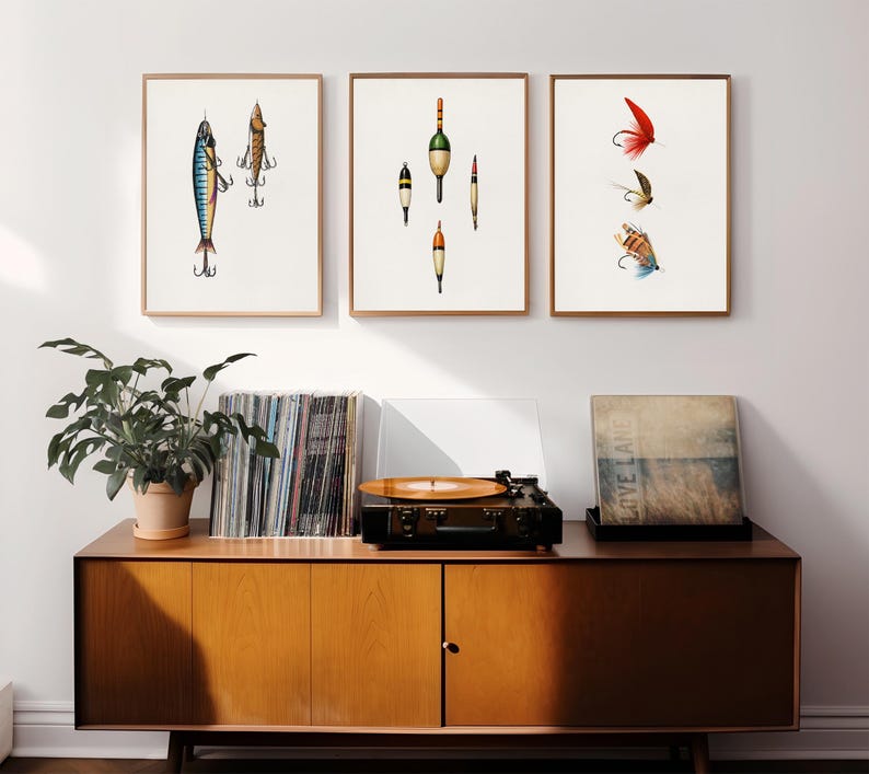 May include: Three framed art prints featuring fishing lures and bobbers. The prints are displayed above a mid-century modern style wooden cabinet with a record player, vinyl records, and a potted plant.