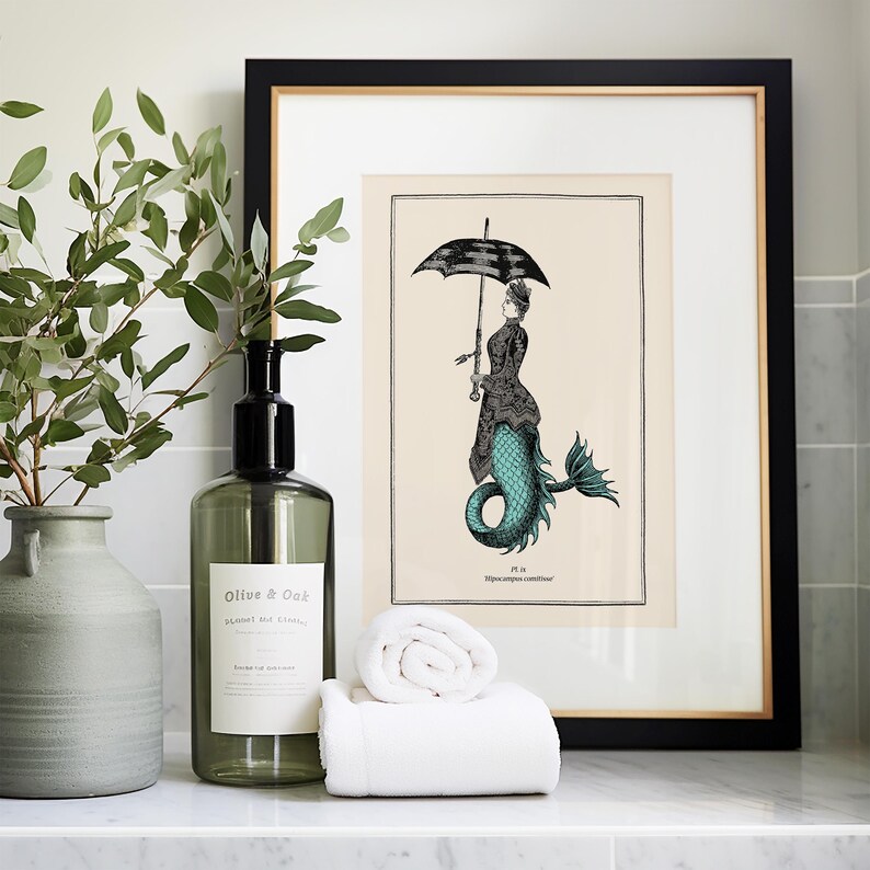 May include: A framed print of a mermaid holding an umbrella. The mermaid is wearing a dress and has a teal tail. The print is on a cream background with black text that reads "Pl. 13. Hippocampus conditor".
