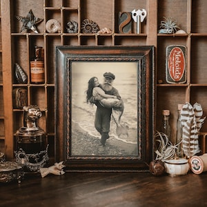 K&ouml;nnte beinhalten: Ein gerahmtes Foto im Vintage-Stil von einem Mann und einer Frau, die einen Meerjungfrauen-Schwanz halten. Das Foto ist vor einem Hintergrund aus Holz mit verschiedenen Dekoartikeln platziert.