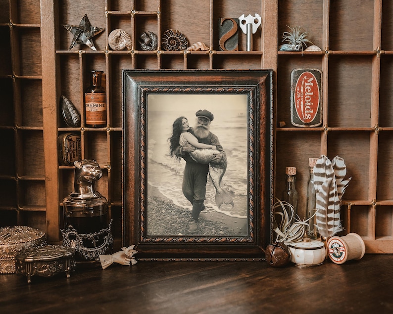 K&ouml;nnte beinhalten: Ein gerahmtes Foto im Vintage-Stil von einem Mann und einer Frau, die einen Meerjungfrauen-Schwanz halten. Das Foto ist vor einem Hintergrund aus Holz mit verschiedenen Dekoartikeln platziert.