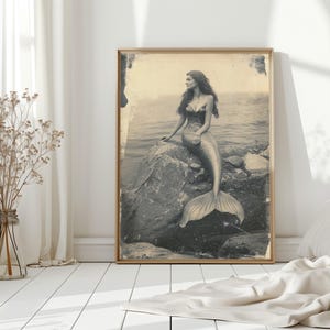May include: A vintage-style photograph of a mermaid sitting on a rock in the ocean. The mermaid has long hair and is wearing a shell bra. The photo is in black and white and has a faded look.