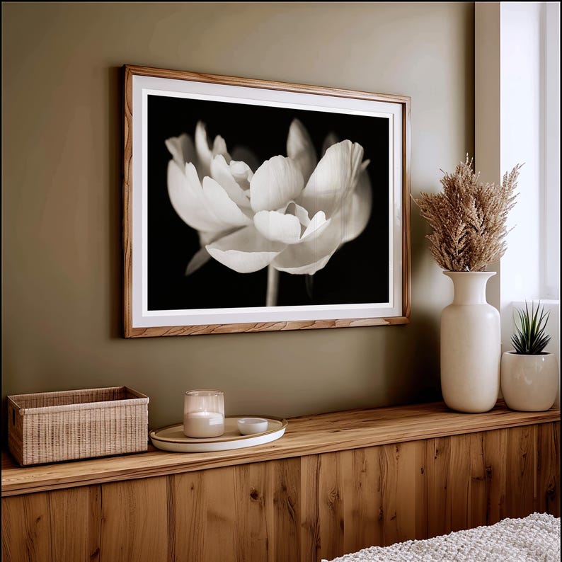 May include: A framed black and white photograph of a flower with delicate petals. The artwork is displayed on a wooden wall, above a wooden shelf with a woven basket, candle, and vase with dried plants. The room has a neutral color palette.