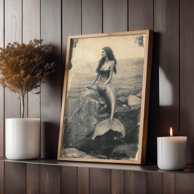 May include: A vintage black and white photograph of a mermaid sitting on a rock. The mermaid has long hair and is wearing a seashell bra. The photo is framed in a wooden frame.