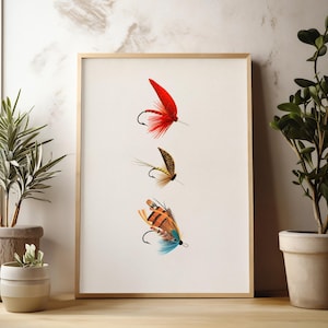 May include: A framed print featuring three colorful fishing flies. The top fly is red with a black hook, the middle fly is brown and tan, and the bottom fly is orange, blue, and brown. The artwork is displayed on a white background.