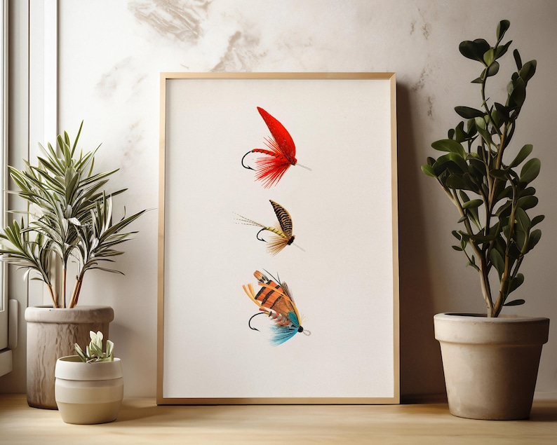 May include: A framed print featuring three colorful fishing flies. The top fly is red with a black hook, the middle fly is brown and tan, and the bottom fly is orange, blue, and brown. The artwork is displayed on a white background.