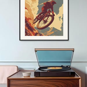 May include: A framed poster with a mountain biking theme. The artwork depicts a cyclist mid-air, with the text "RIDE, CRASH, SWEAR, REPEAT." The poster is displayed above a record player on a wooden cabinet.