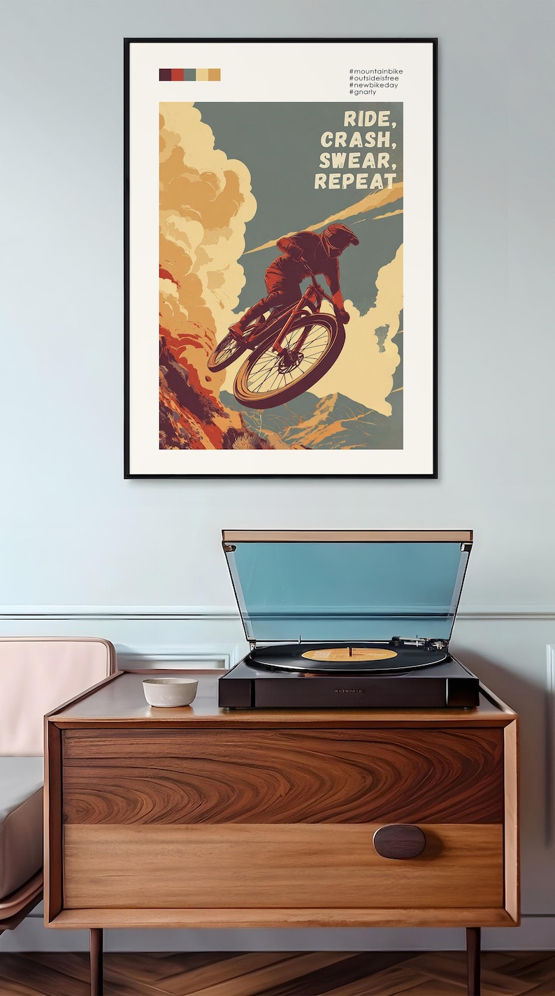May include: A framed poster with a mountain biking theme. The artwork depicts a cyclist mid-air, with the text "RIDE, CRASH, SWEAR, REPEAT." The poster is displayed above a record player on a wooden cabinet.