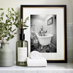 May include: A black and white photograph of a robot taking a bath in a white bathtub. The photograph is framed in a black frame with a gold trim. The photograph is sitting on a white countertop with a green glass bottle, a green ceramic vase, and a white towel.