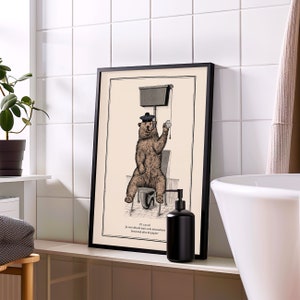 May include: A black framed print of a bear wearing a beret sitting on a toilet. The print is in a bathroom setting with a white tiled wall, a white sink, and a black soap dispenser. The text on the print reads "Je suis d&eacute;sol&eacute; mais cela n&eacute;cessitera beaucoup plus de papier."