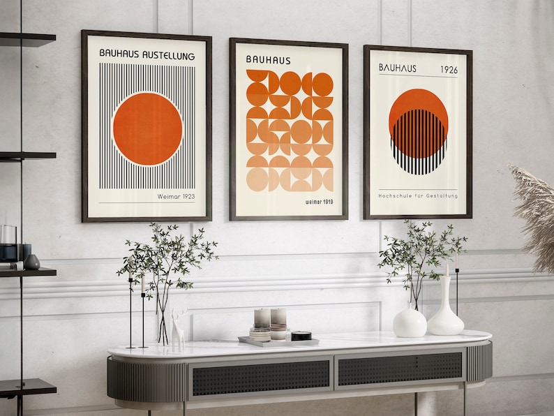 May include: Three framed prints with geometric designs in black, white, and orange. The prints feature the text "Bauhaus" and the years "1923", "1919", and "1926".