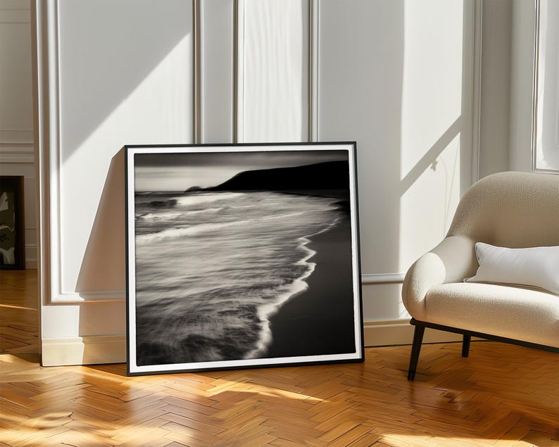 May include: Black and white photograph of a beach with waves crashing on the shore. The photo is framed in a black frame.