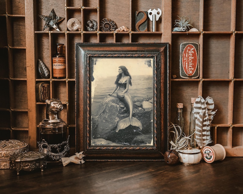 May include: A vintage framed photograph of a mermaid with long flowing hair, sitting on a rock in the ocean. The photo is in a dark wood frame with a rope design.