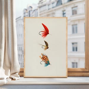 May include: A framed print featuring three detailed fishing flies. The flies are in shades of red, brown, orange, and blue, with curved hooks. The background is a cream color, and the frame is light wood.