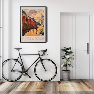 May include: A vintage poster advertising the Vuelta a Espana cycling race. The poster features a scenic mountain road with cyclists riding up a steep incline. The poster is framed and hanging on a white wall.