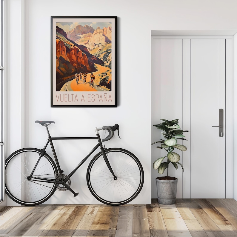 May include: A vintage poster advertising the Vuelta a Espana cycling race. The poster features a scenic mountain road with cyclists riding up a steep incline. The poster is framed and hanging on a white wall.
