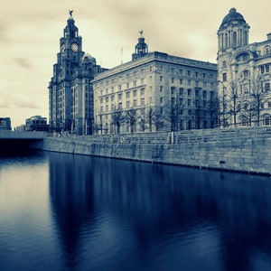 May include: A sepia-toned photograph of the Liverpool waterfront featuring several large, ornate buildings with clock towers and domes. The buildings are reflected in the calm water of the river, creating a symmetrical image.