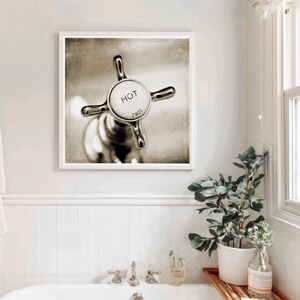 May include: A sepia-toned framed print of a vintage faucet handle. The handle is chrome with a white center that reads "HOT" and "1901". The image is in a bathroom setting with a white tub and a plant.