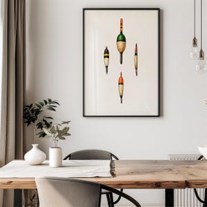 May include: A framed print featuring four vintage fishing bobbers in various shapes and colors. The bobbers are painted with orange, green, yellow, and black accents against a cream-colored background. The artwork is displayed in a black frame.
