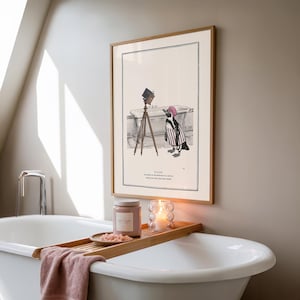 May include: A framed illustration depicts a penguin in a striped outfit and a pink hat, standing near a bathtub with a vintage camera on a tripod. A wooden tray holds a candle, a jar, and a small dish with pink contents, placed over a white bathtub.