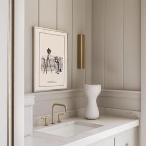 May include: A framed illustration depicts a penguin photographer with a camera on a tripod. The penguin is dressed in a striped outfit and a pink hat. The artwork is displayed above a white sink with gold fixtures, a white vase, and a gold wall sconce.