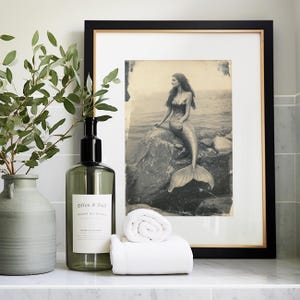 May include: A framed black and white photo of a mermaid sitting on a rock in the water. The photo is in a black frame with a gold trim. There is a green glass bottle with a pump dispenser and a white towel rolled up in front of the frame.