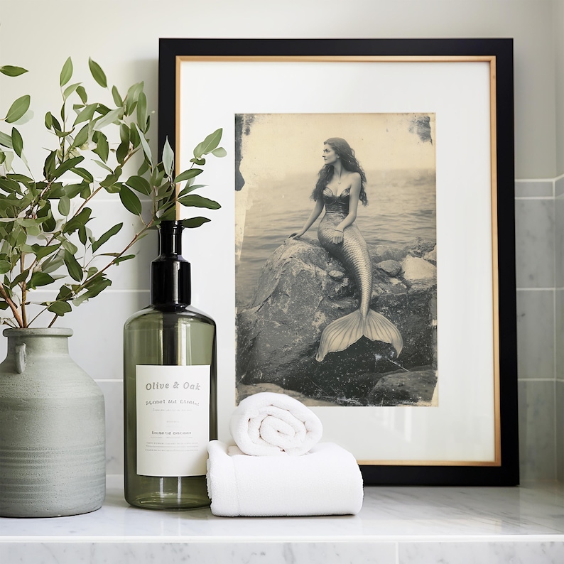 May include: A framed black and white photo of a mermaid sitting on a rock in the water. The photo is in a black frame with a gold trim. There is a green glass bottle with a pump dispenser and a white towel rolled up in front of the frame.