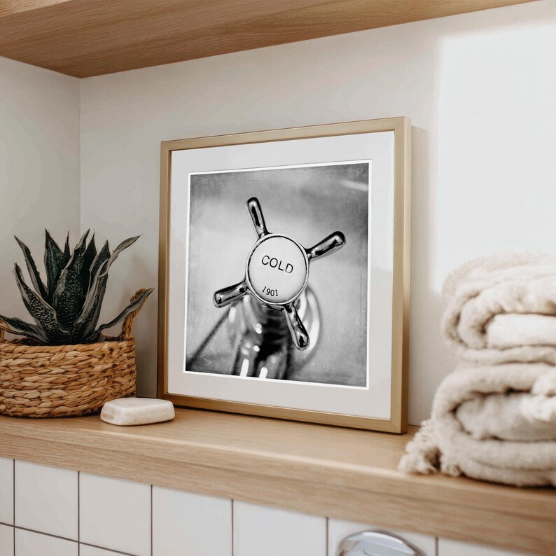 May include: Black and white framed print of a vintage faucet handle. The handle is chrome with a white center that reads "COLD" and "1901". The print is displayed on a wooden shelf with a woven basket, a bar of soap, and a stack of towels.
