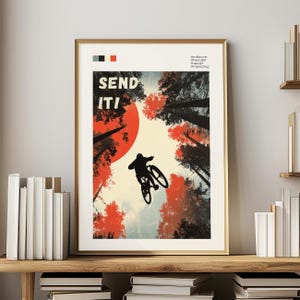 May include: A framed poster with a gold border features a mountain biker silhouetted against a red sun and tall trees. The words "SEND IT!" are in bold letters. The color palette includes red, black, and cream.