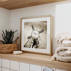 May include: Framed black and white photograph of a vintage faucet handle. The handle is chrome with a white center that reads "COLD" and "1901". The photograph is in a gold frame and is displayed on a wooden shelf with a woven basket and towels.